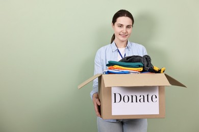 Photo of Woman holding donation box with clothes on pale olive background, space for text