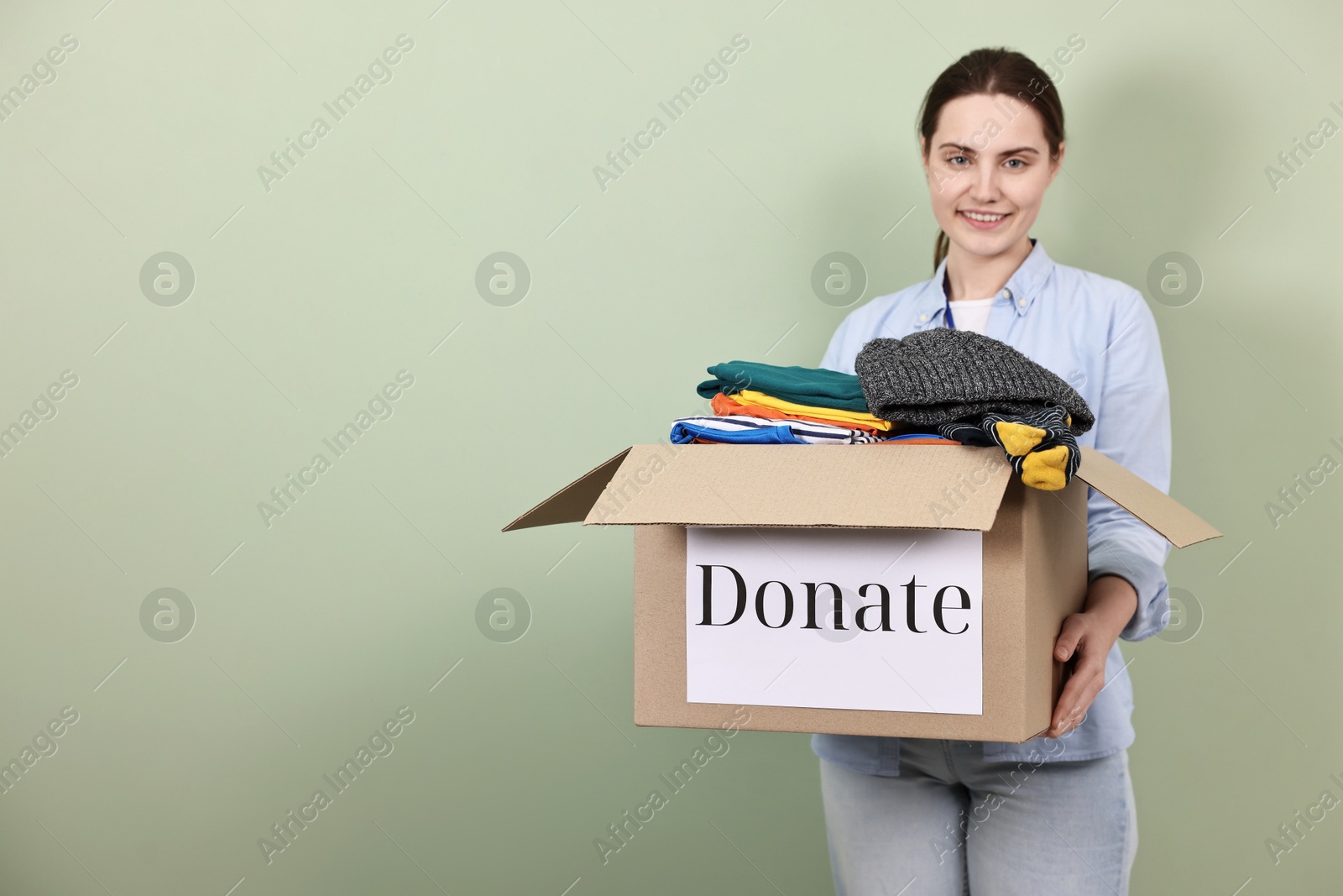 Photo of Woman holding donation box with clothes on pale olive background, space for text