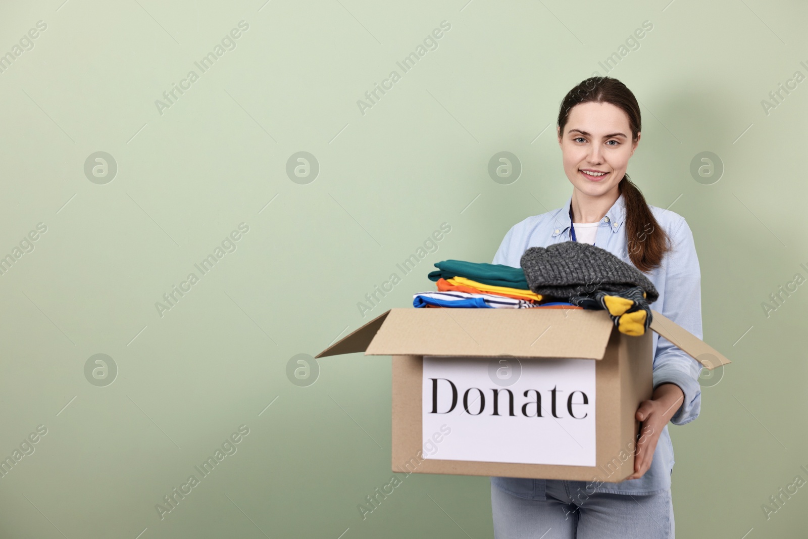 Photo of Woman holding donation box with clothes on pale olive background, space for text