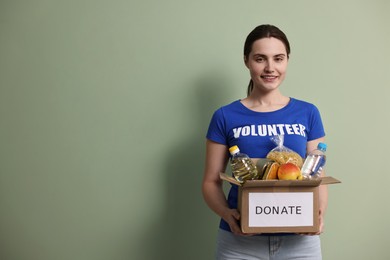 Photo of Woman holding donation box with food products on pale olive background, space for text