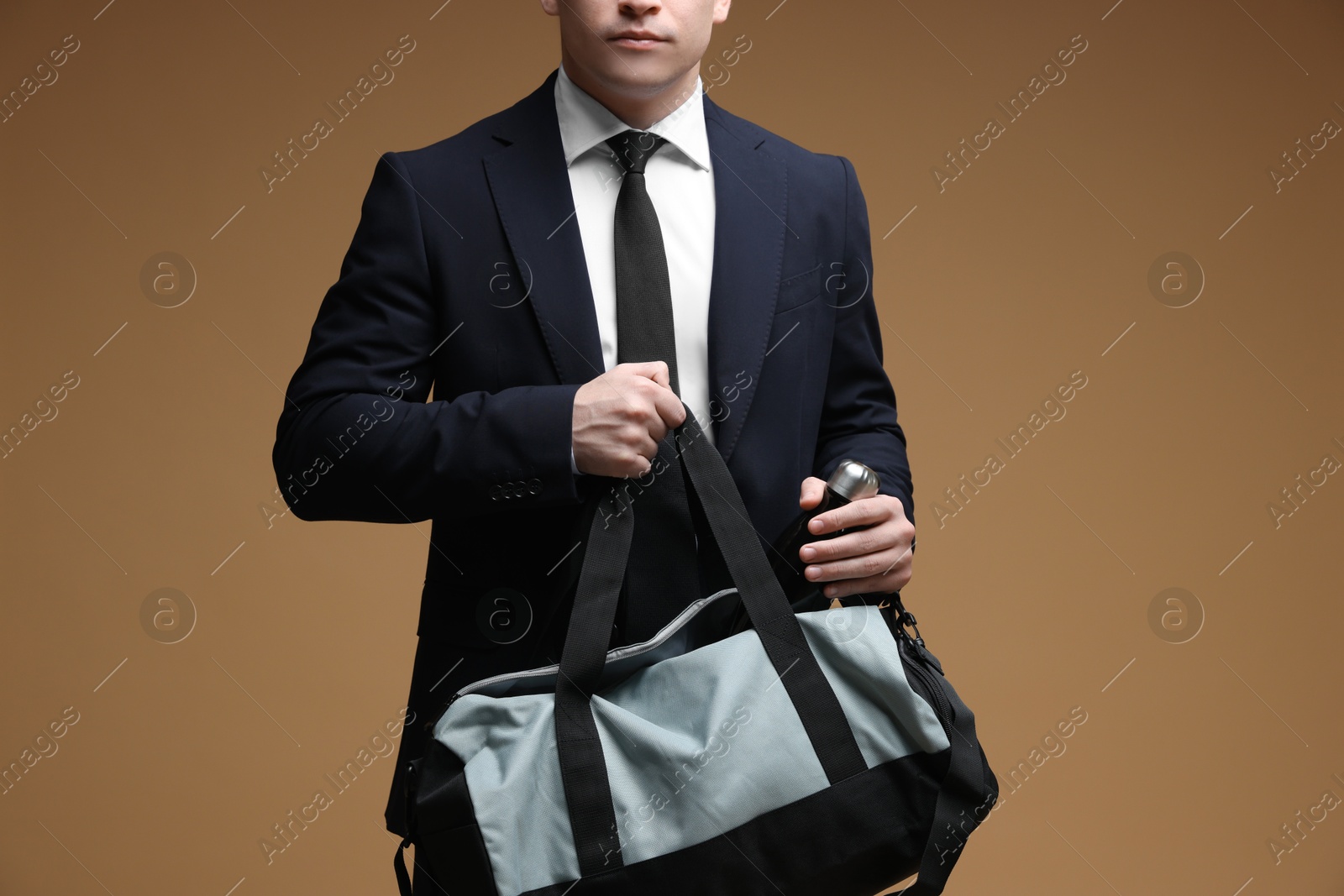 Photo of Man in suit putting bottle of water into gym bag on brown background, closeup