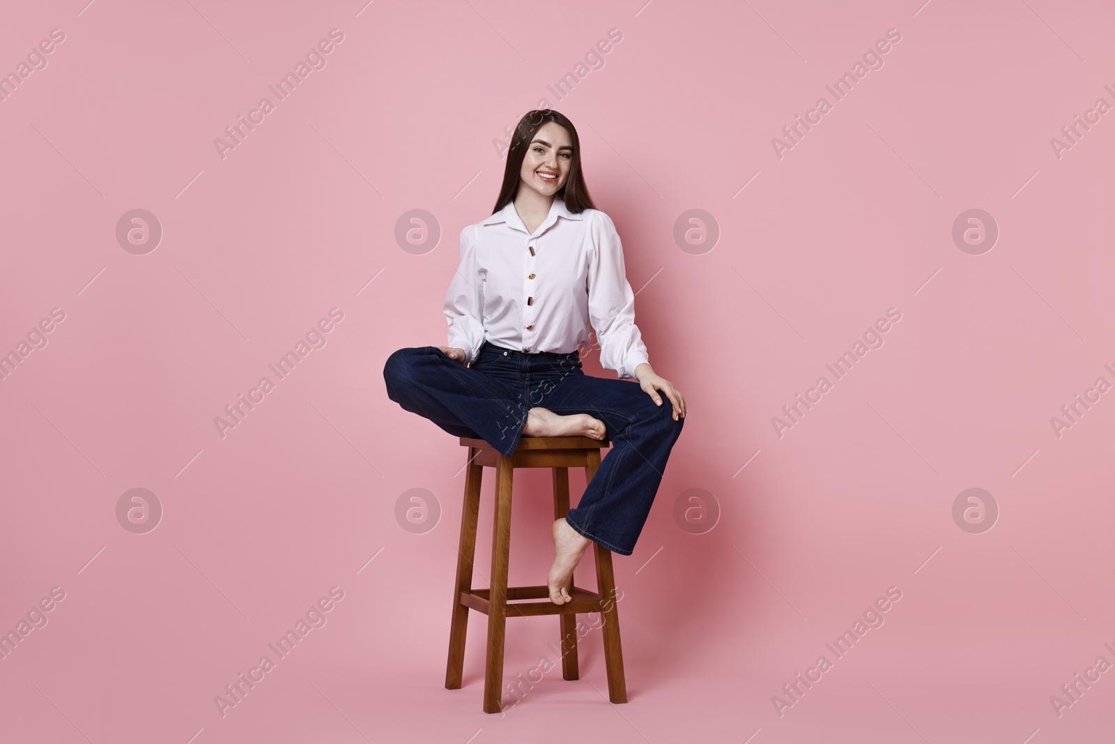 Photo of Smiling woman in stylish jeans sitting on stool against pink background