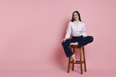 Smiling woman in stylish jeans sitting on stool against pink background. Space for text