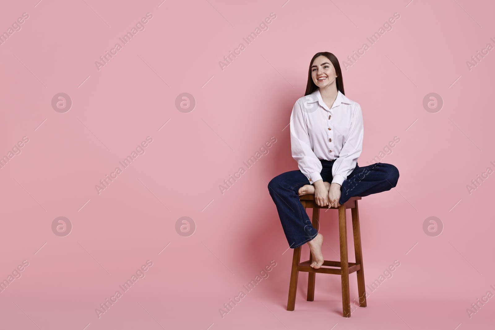 Photo of Smiling woman in stylish jeans sitting on stool against pink background. Space for text