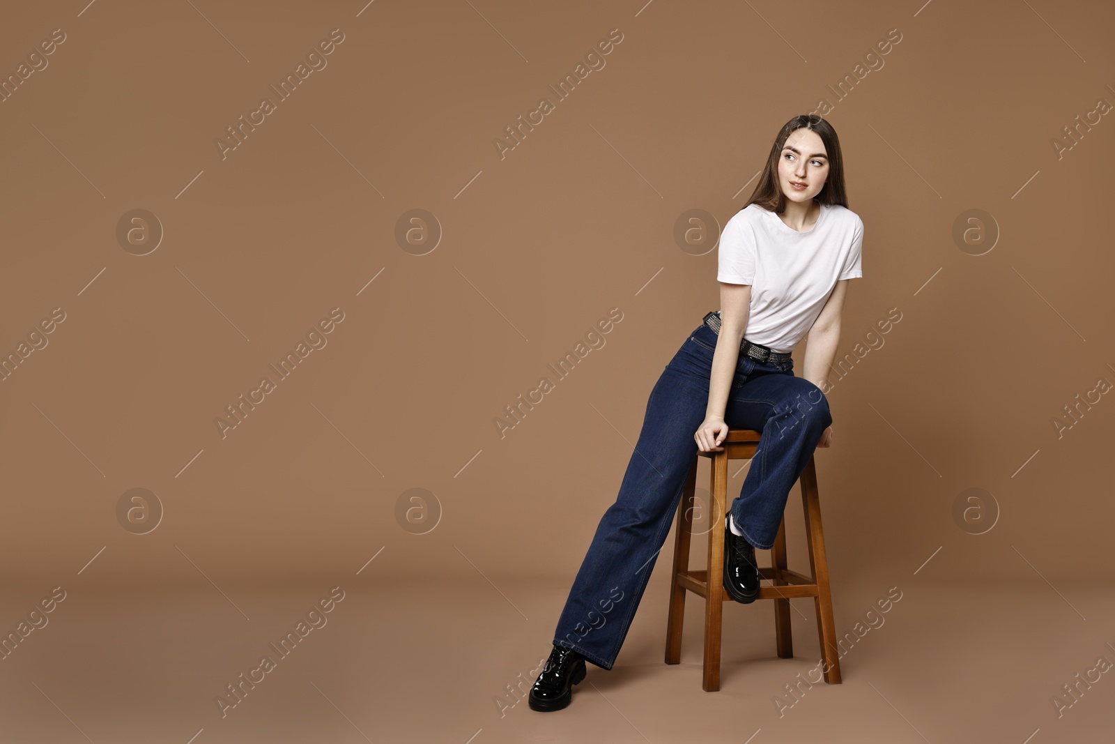 Photo of Woman in stylish jeans sitting on stool against brown background. Space for text