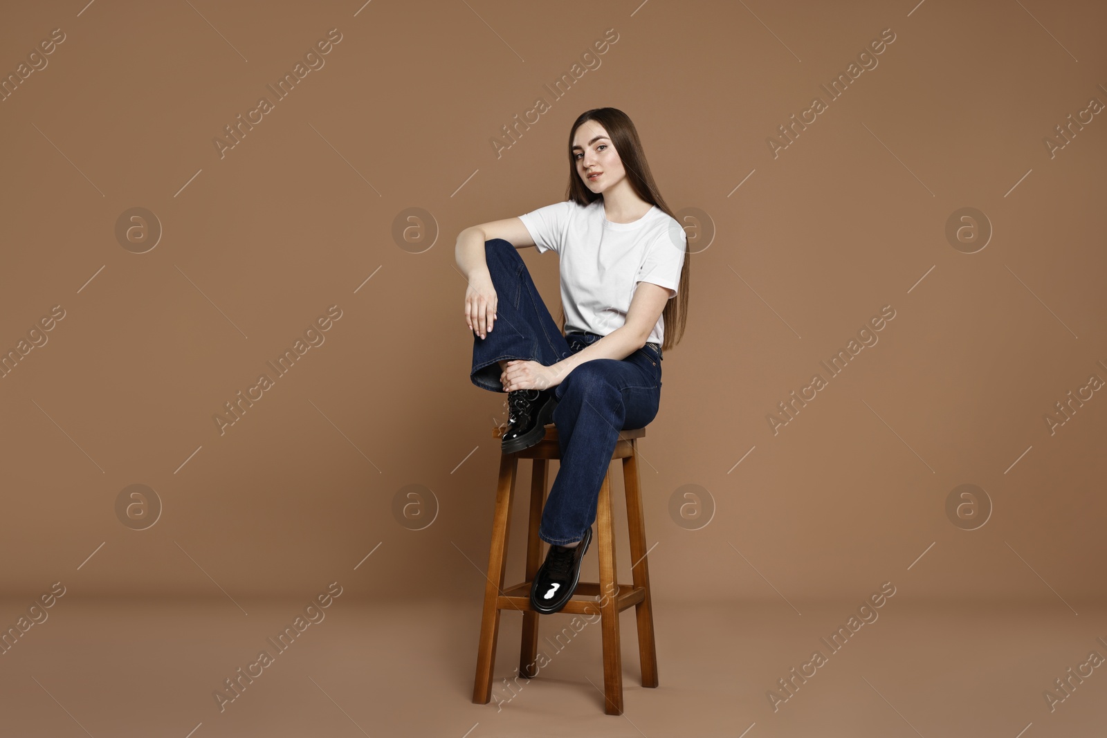 Photo of Beautiful young woman in stylish jeans sitting on stool against brown background