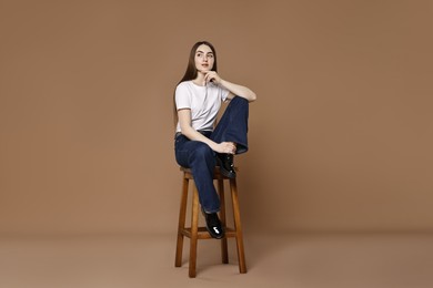 Photo of Beautiful young woman in stylish jeans sitting on stool against brown background