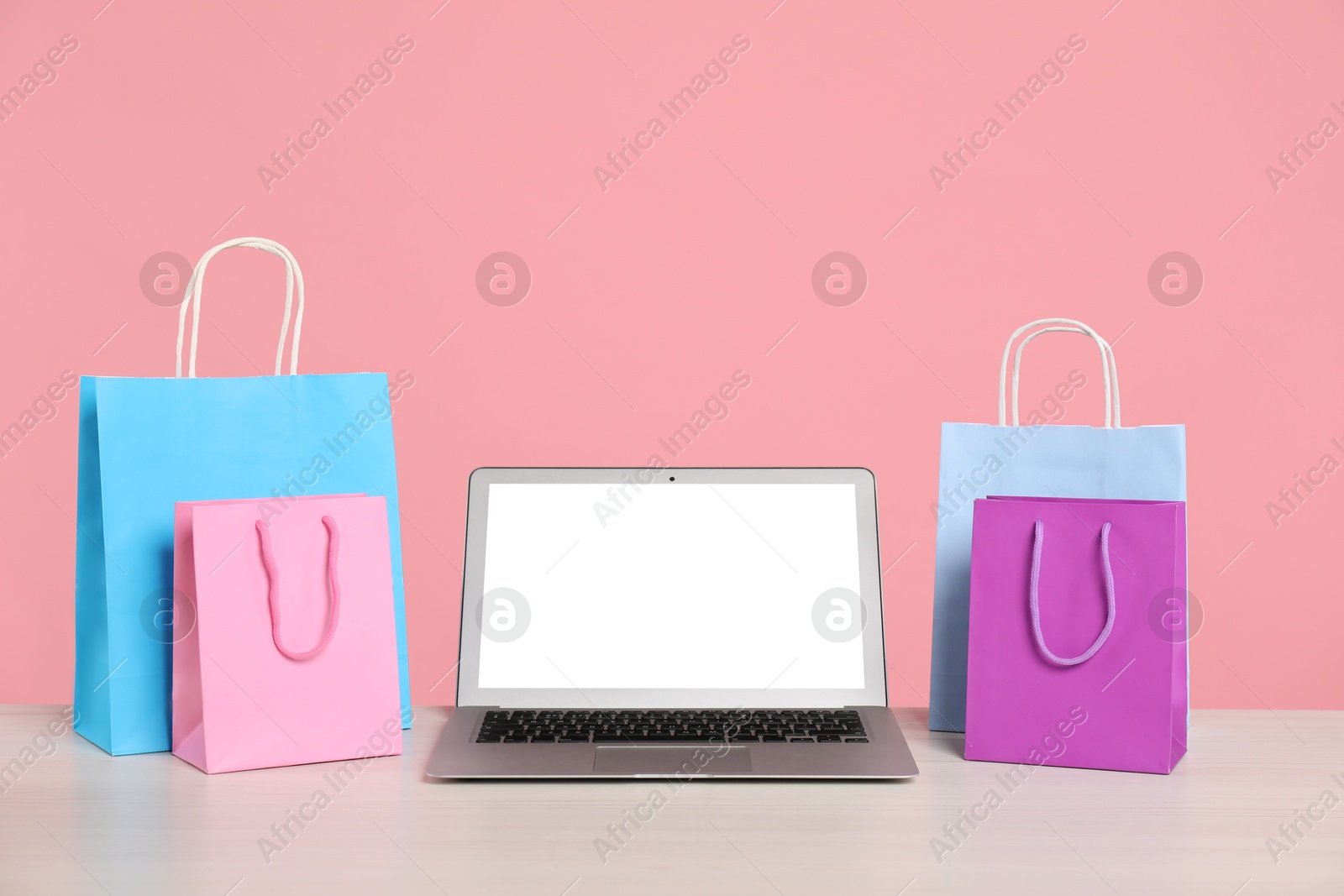 Photo of Internet shopping. Laptop and colorful paper bags on table against pink background