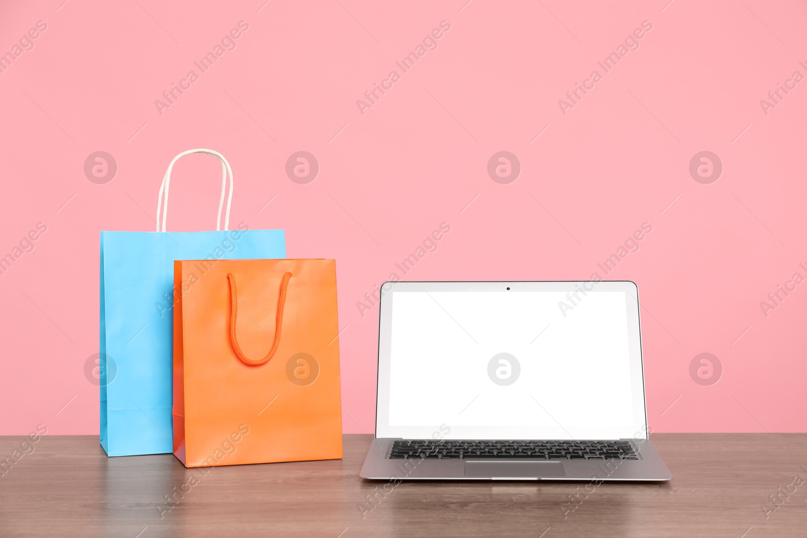 Photo of Internet shopping. Laptop and colorful paper bags on wooden table against pink background