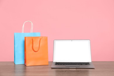 Photo of Internet shopping. Laptop and colorful paper bags on wooden table against pink background