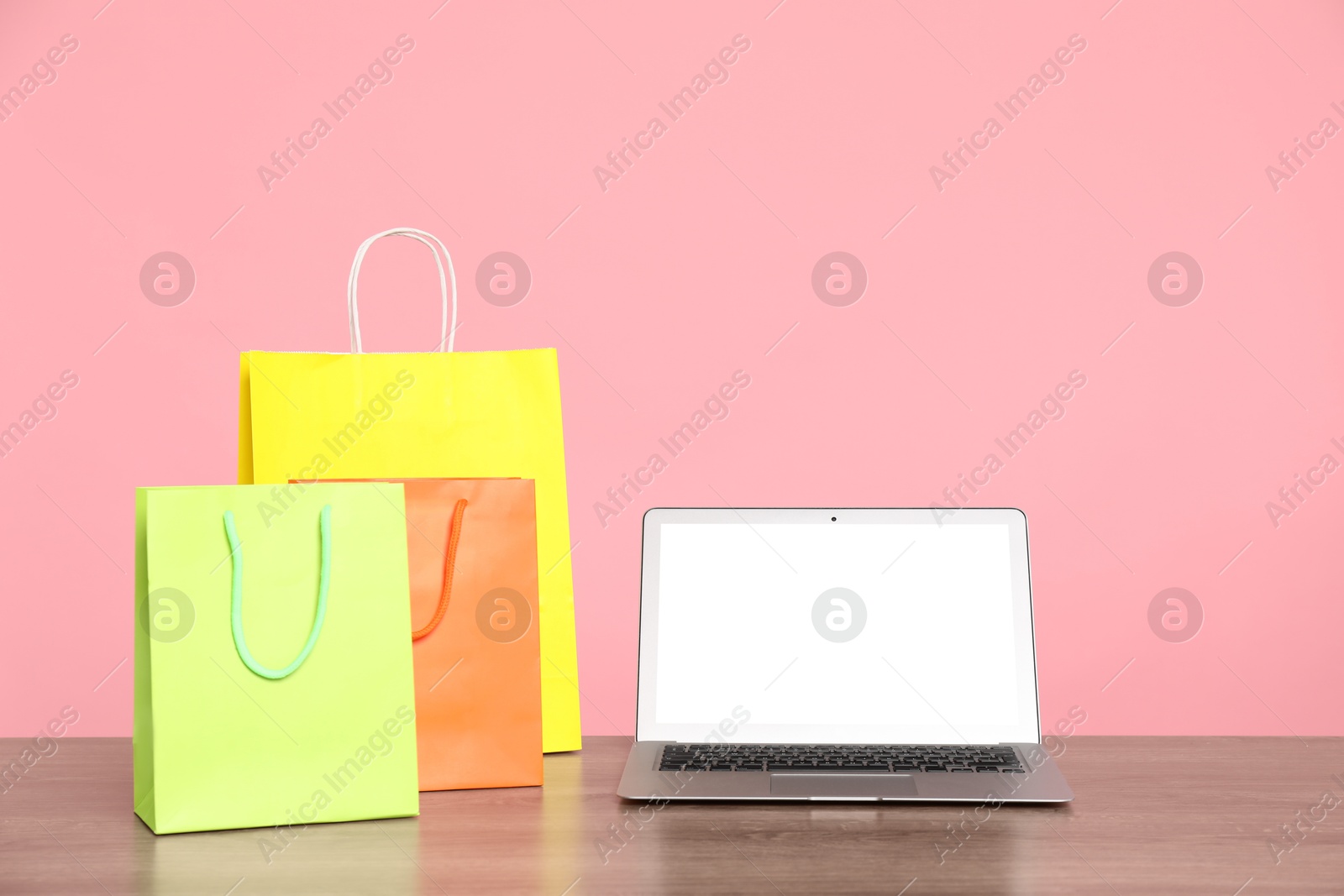 Photo of Internet shopping. Laptop and colorful paper bags on wooden table against pink background