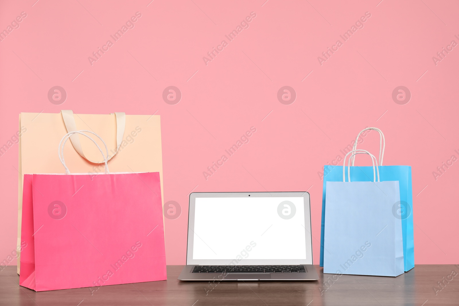 Photo of Internet shopping. Laptop and colorful paper bags on wooden table against pink background