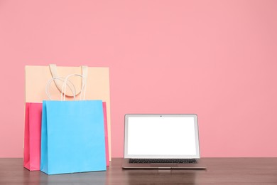 Photo of Internet shopping. Laptop and colorful paper bags on wooden table against pink background