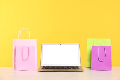 Photo of Internet shopping. Laptop and colorful paper bags on table against yellow background