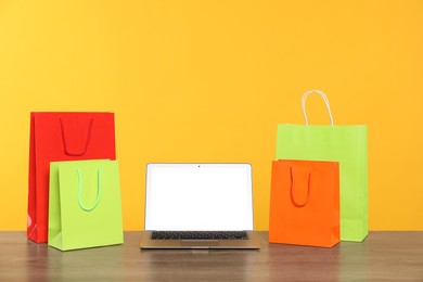 Photo of Internet shopping. Laptop and colorful paper bags on wooden table against yellow background