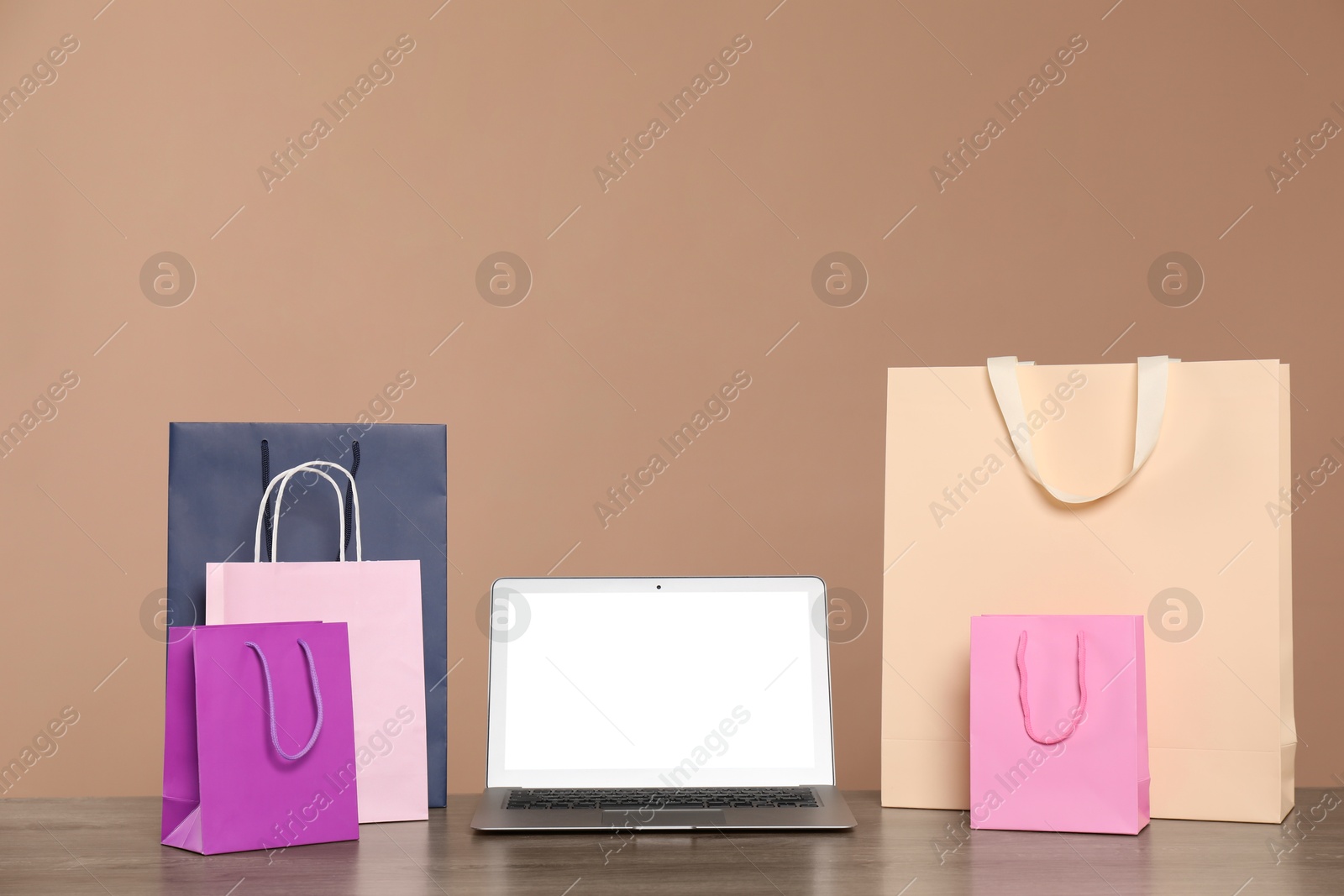 Photo of Internet shopping. Laptop and colorful paper bags on wooden table against brown background