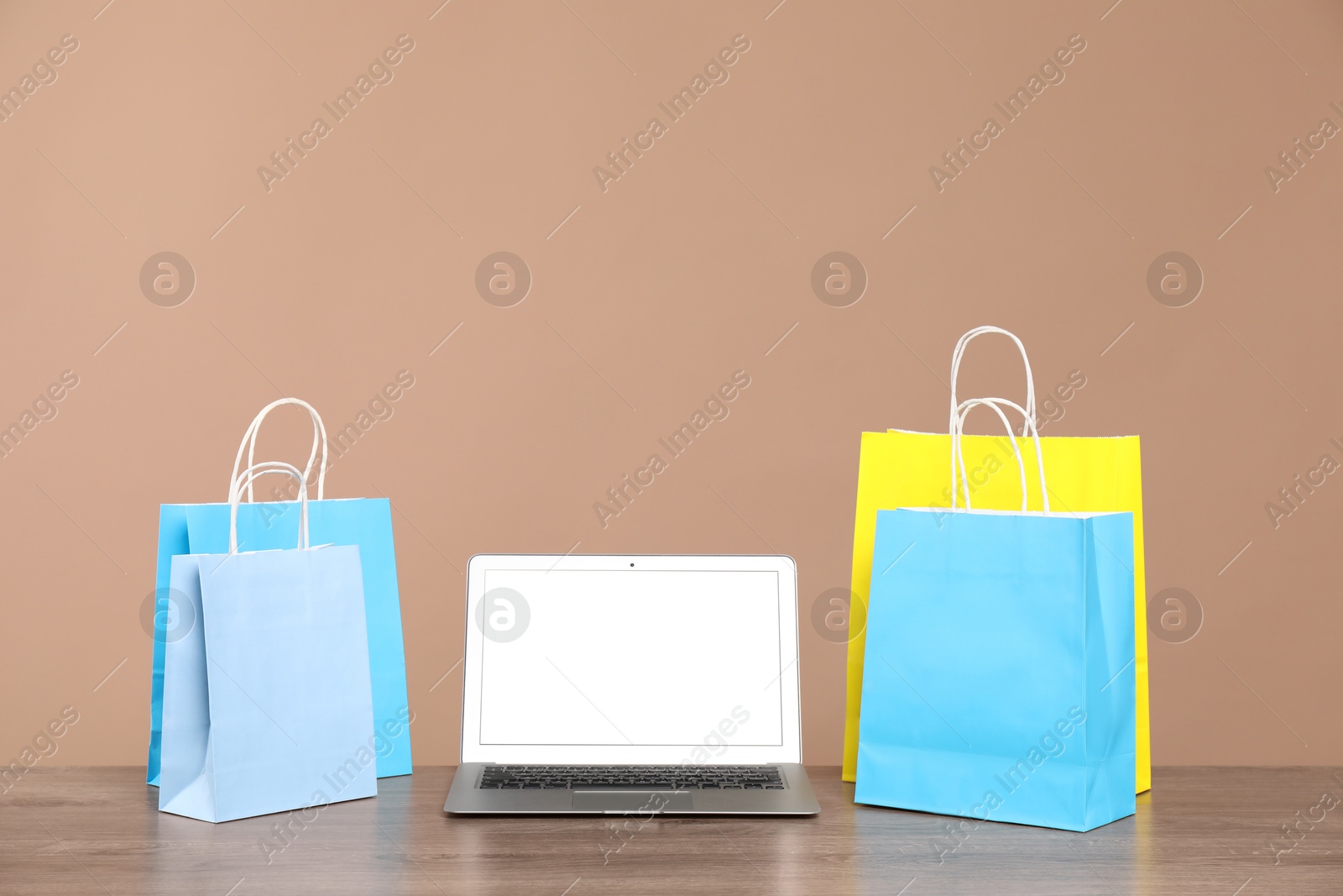 Photo of Internet shopping. Laptop and colorful paper bags on wooden table against brown background