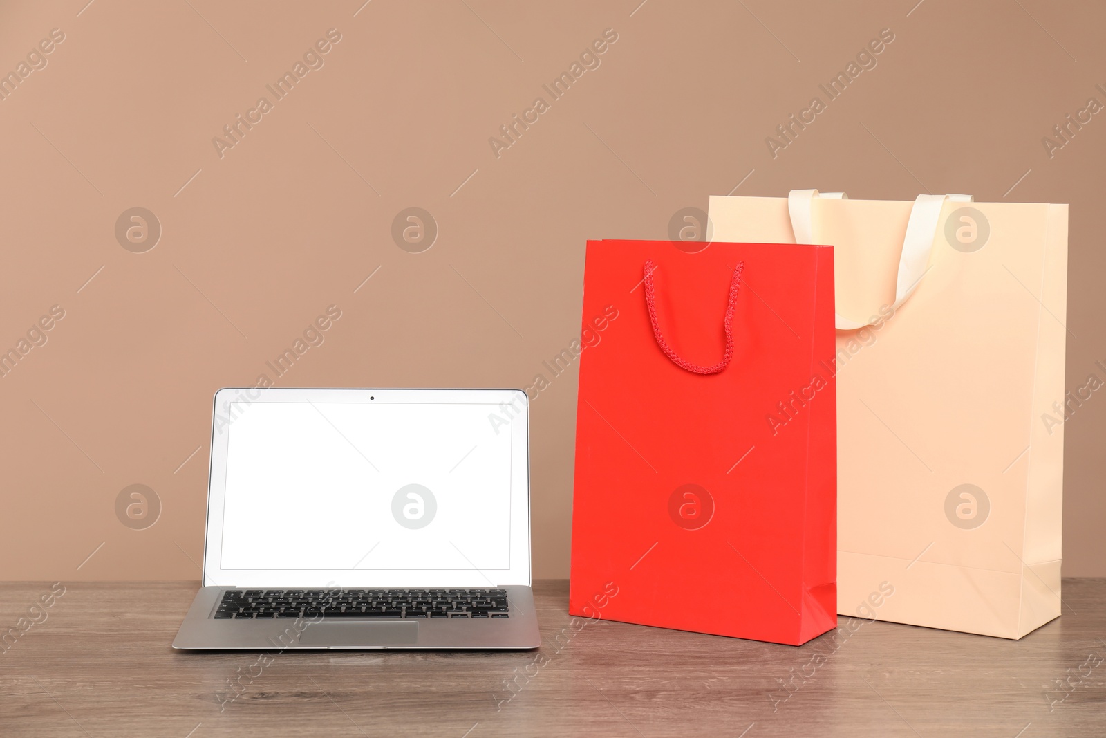 Photo of Internet shopping. Laptop and colorful paper bags on wooden table against brown background