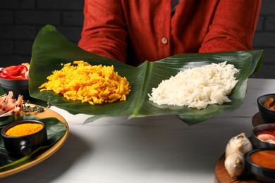 Photo of Woman holding cut banana leaf with piles of rice at white table, closeup. Healthy eco serving