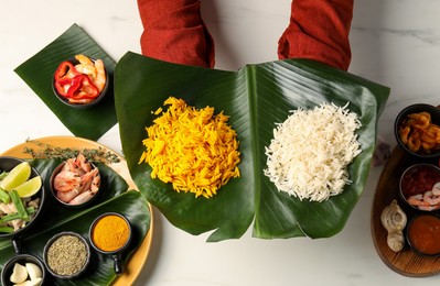 Photo of Woman holding cut banana leaf with piles of rice at white table, top view. Healthy eco serving