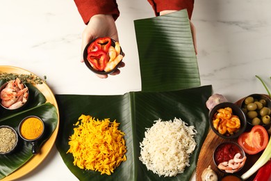 Photo of Woman holding piece of banana leaf and bowl with shrimps at white marble table, top view