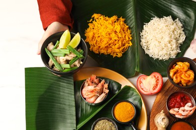 Photo of Woman holding bowl with tasty food at white table with pieces of banana leaves, top view