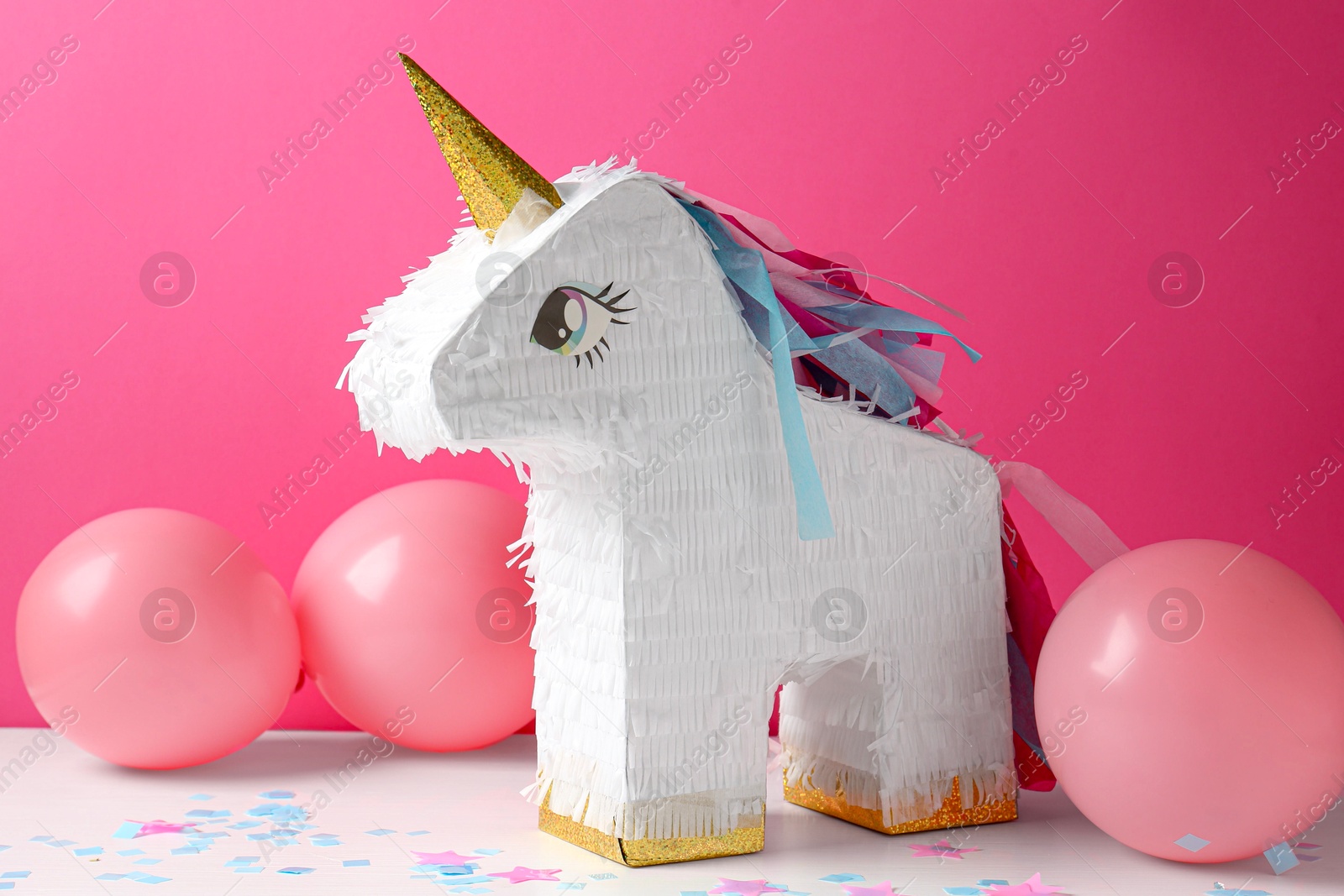 Photo of Bright pinata in shape of unicorn and balloons on white table against pink background