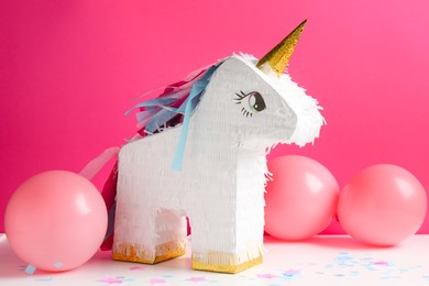 Photo of Bright pinata in shape of unicorn and balloons on white table against pink background