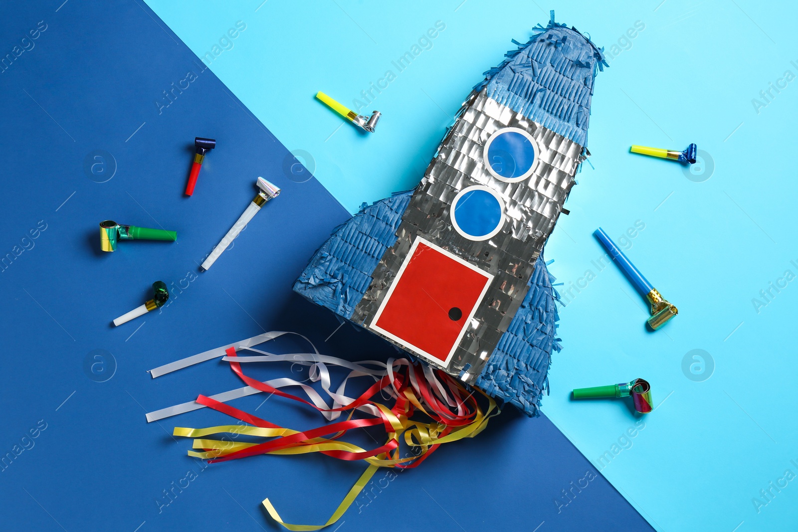 Photo of Bright pinata in shape of spaceship and blowers on blue background, flat lay