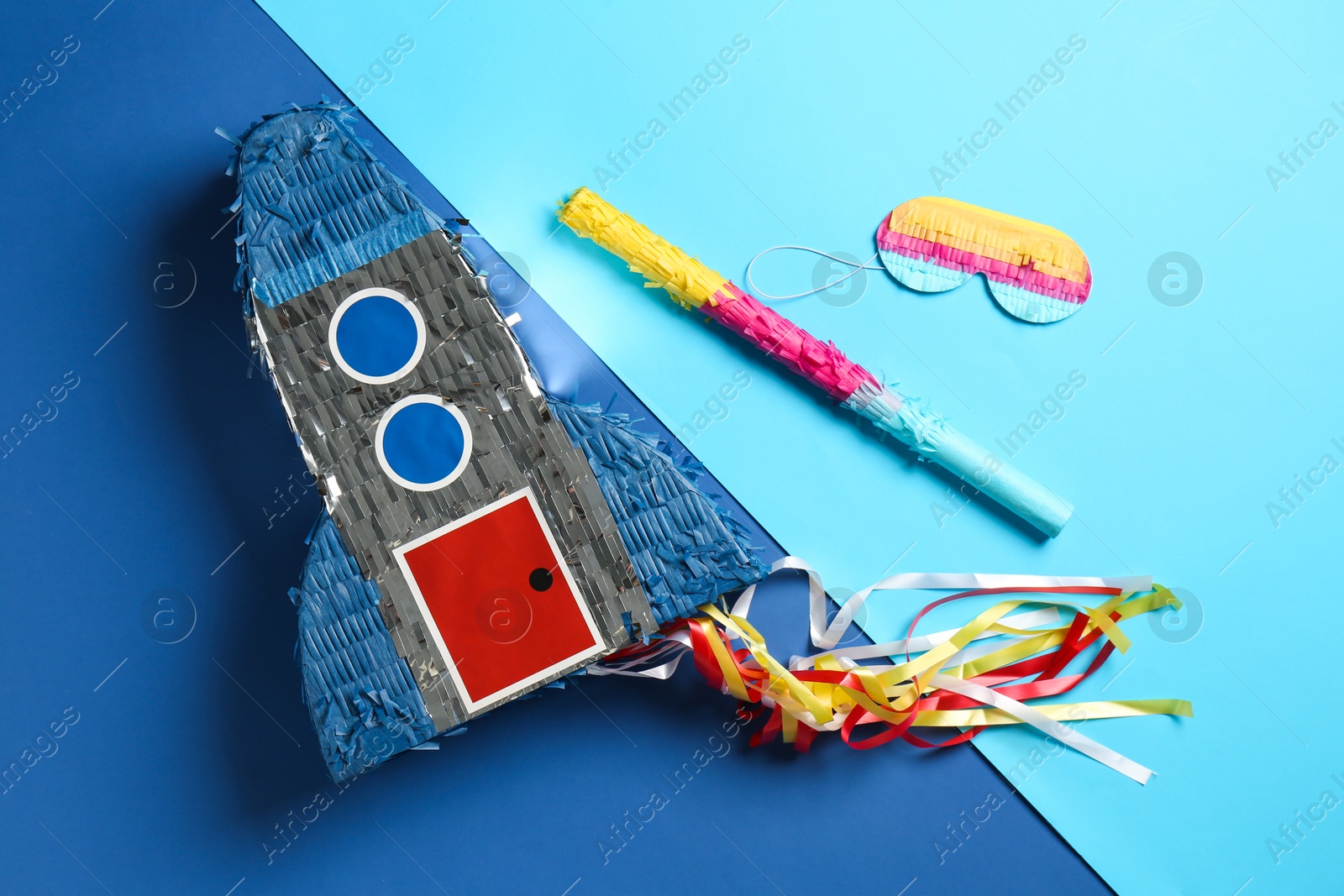 Photo of Bright pinata in shape of spaceship , blindfold and stick on blue background, flat lay