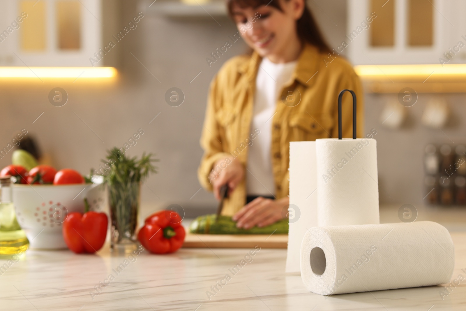 Photo of Woman cutting zucchini at white marble table in kitchen, focus on rolls of paper towels