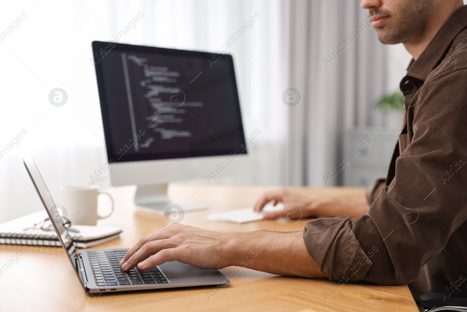 Photo of Programmer working on laptop and computer at wooden desk indoors, closeup