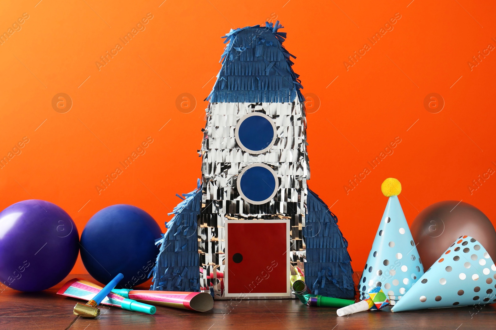 Photo of Bright pinata in shape of spaceship and party accessories on table against orange background