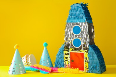 Photo of Bright pinata in shape of spaceship, stick and party hats on yellow background. Space for text