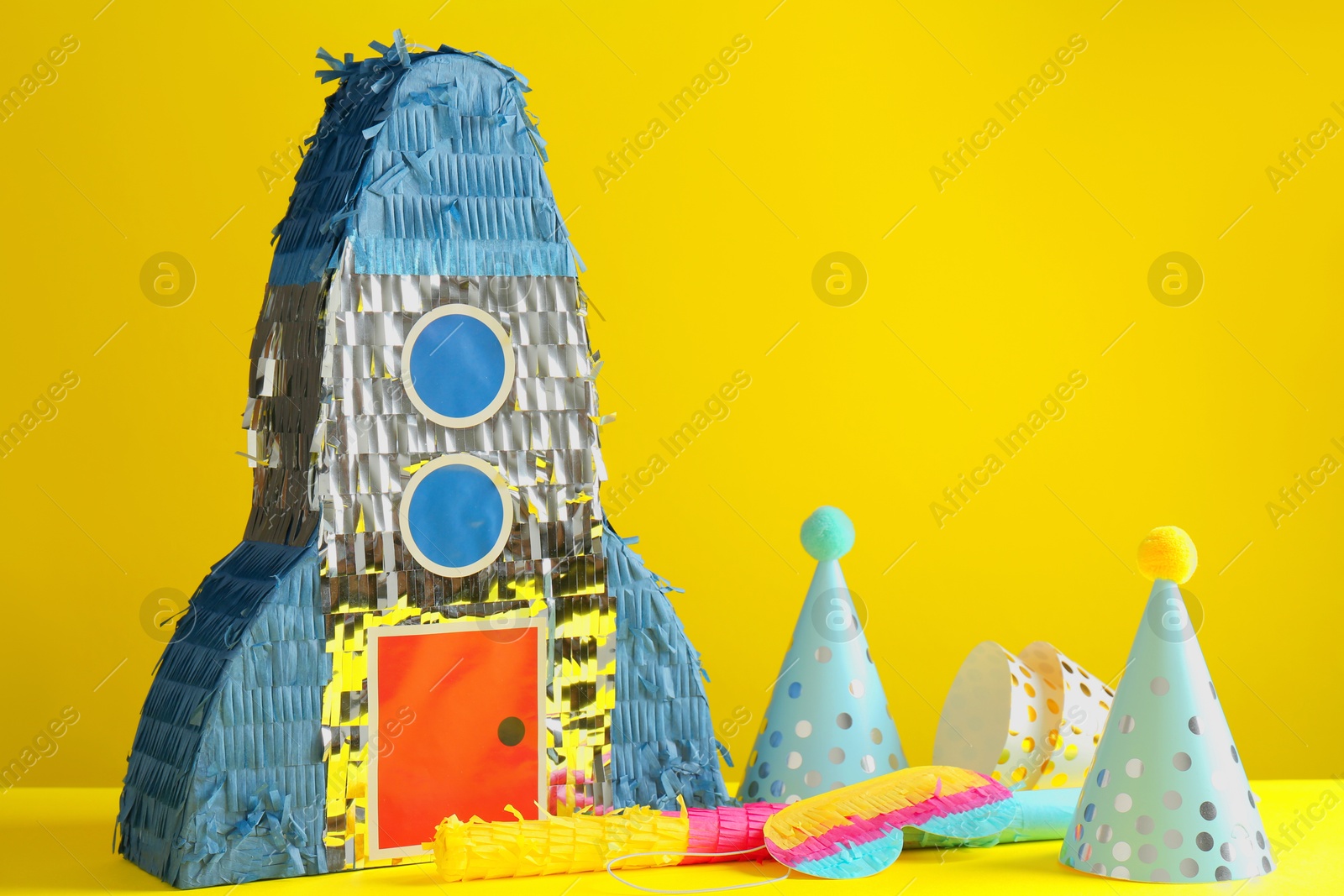 Photo of Bright pinata in shape of spaceship, stick and party hats on yellow background. Space for text