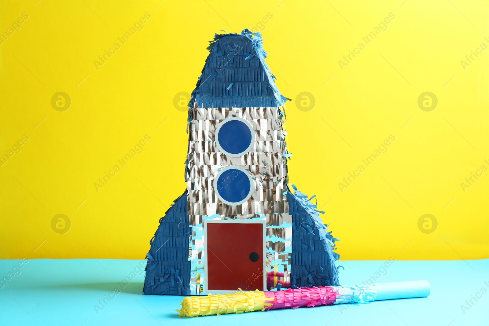 Photo of Beautiful pinata in shape of rocket and stick on blue table against yellow background