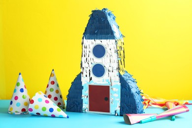 Photo of Beautiful pinata in shape of rocket, party cones and poppers on blue table against yellow background