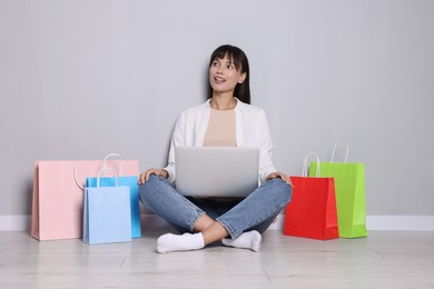 Photo of Internet shopping. Happy woman with laptop and colorful bags sitting near grey wall