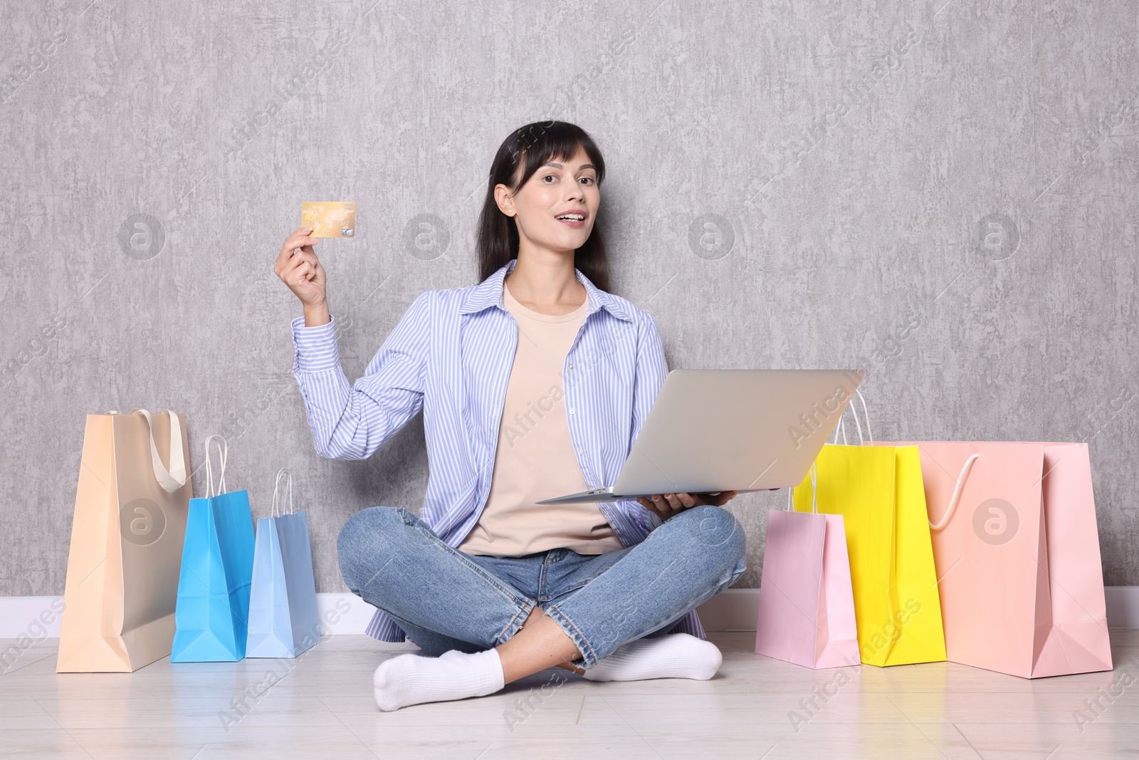 Photo of Internet shopping. Happy woman with credit card, laptop and colorful bags sitting near grey wall