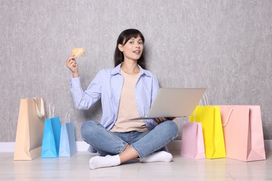 Photo of Internet shopping. Happy woman with credit card, laptop and colorful bags sitting near grey wall