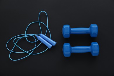 Photo of Dumbbells and skipping rope on black background, flat lay. Sports equipment