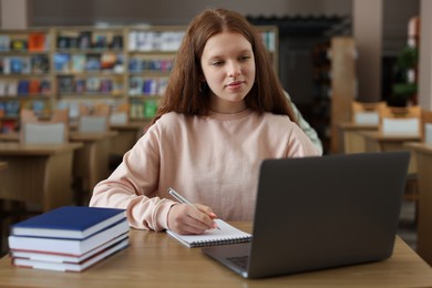 Photo of Girl taking notes while using laptop at wooden desk in public library