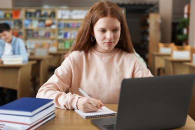 Photo of Girl taking notes while using laptop at wooden desk in public library