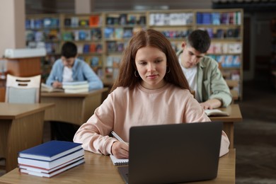 Girl taking notes while using laptop at wooden desk in public library