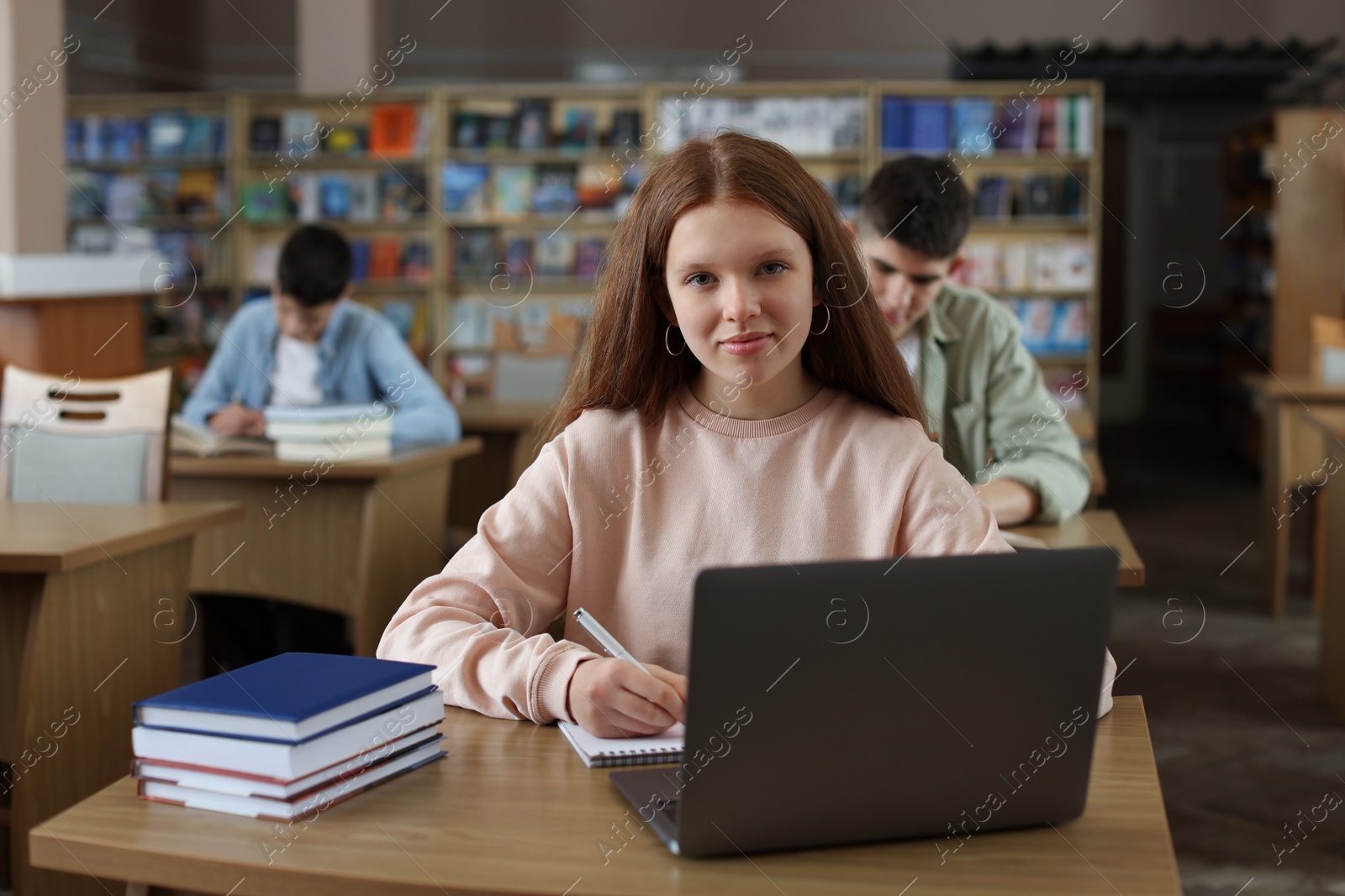 Photo of Girl taking notes while using laptop at wooden desk in public library