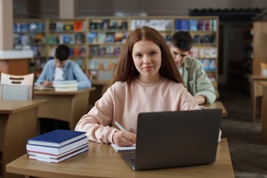 Photo of Girl taking notes while using laptop at wooden desk in public library