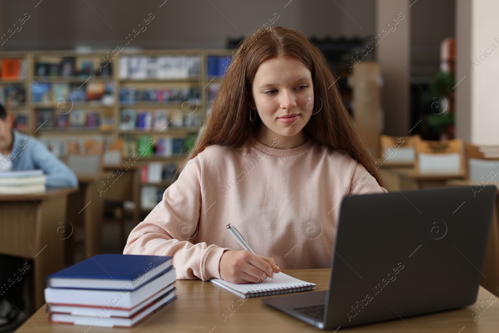 Photo of Girl taking notes while using laptop at wooden desk in public library