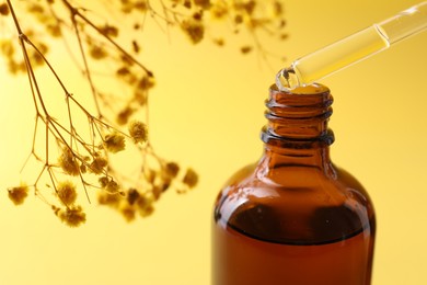 Photo of Dripping serum from pipette into bottle and gypsophila flowers on yellow background, closeup