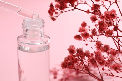 Photo of Dripping serum from pipette into bottle and gypsophila flowers on pink background, closeup