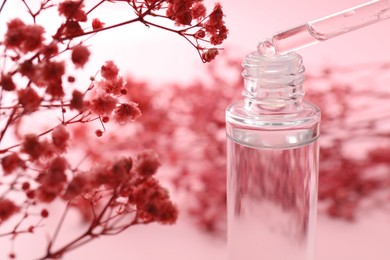 Photo of Dripping serum from pipette into bottle and gypsophila flowers on pink background, closeup